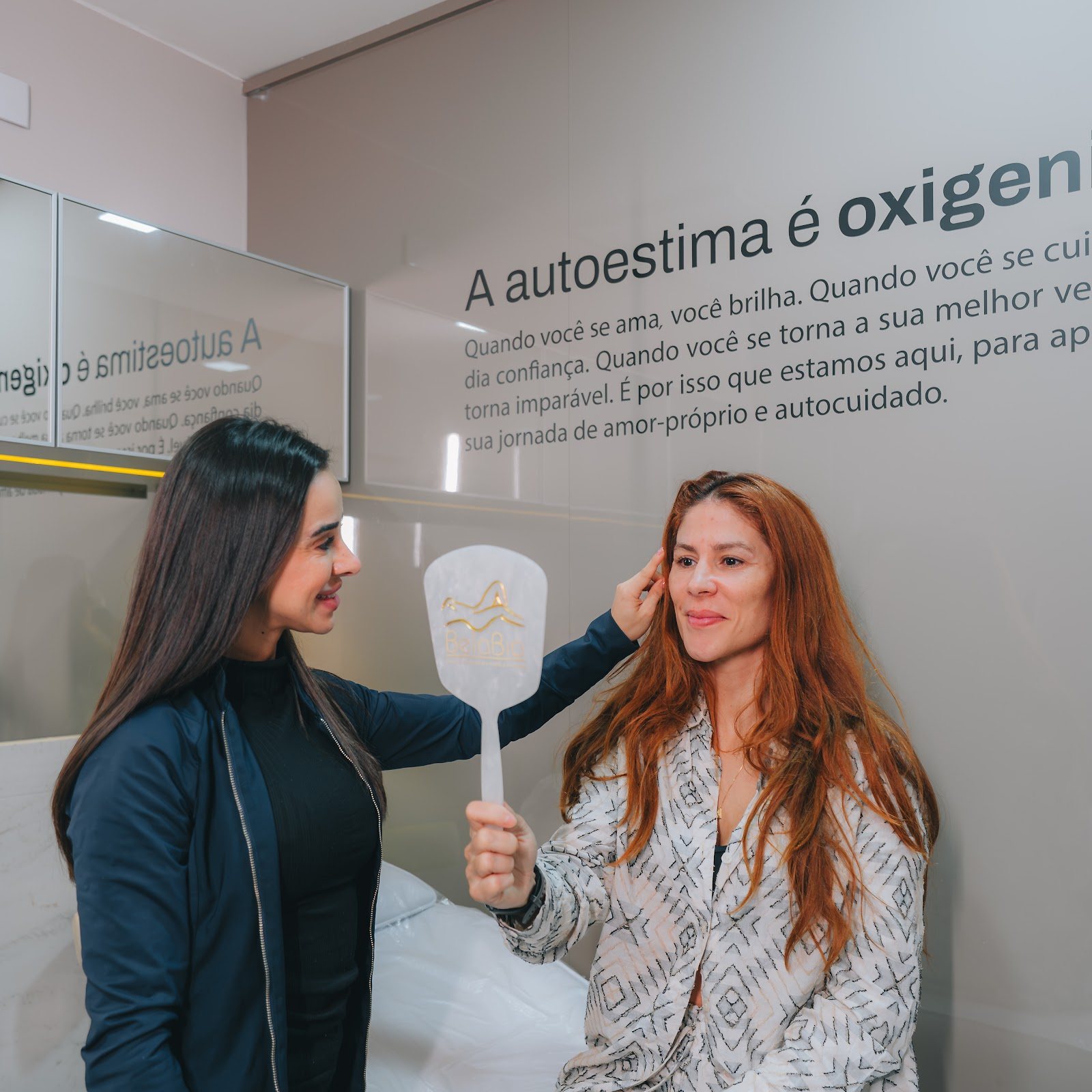 Sala de atendimento da BelaBia: profissional mostra o espelho de mão para uma cliente; na parede, o texto A autoestima é oxigênio